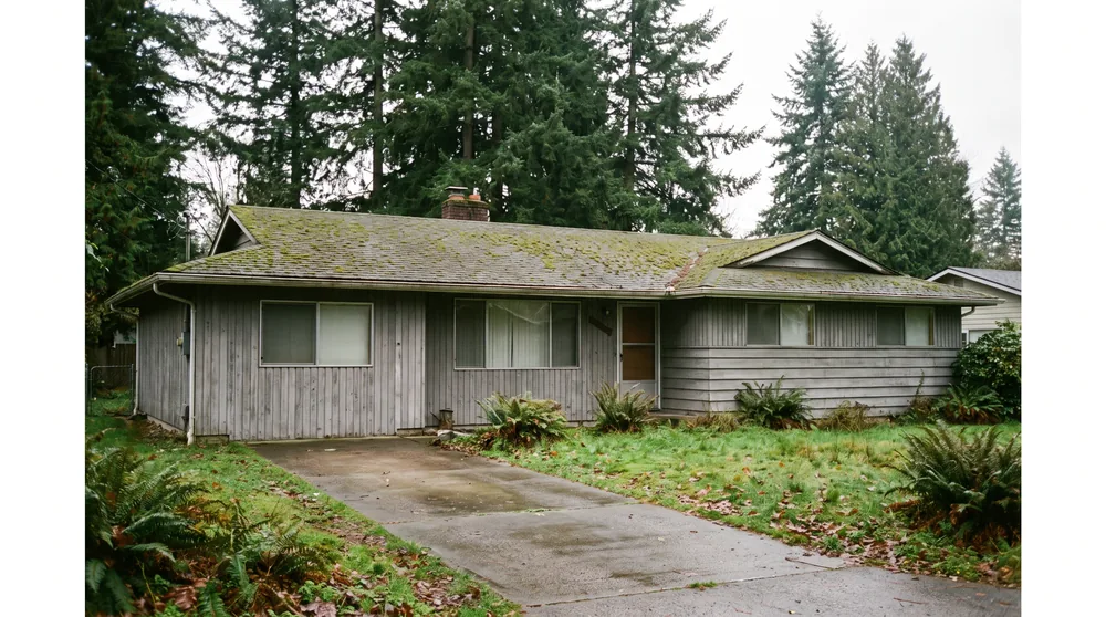 Illustration: a modest gray rambler with a mossy roof and a wet driveway under tall firs, as it stands today