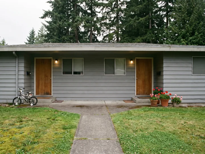 Illustration: a lived-in gray duplex with two front doors, a bike by one and geraniums by the other